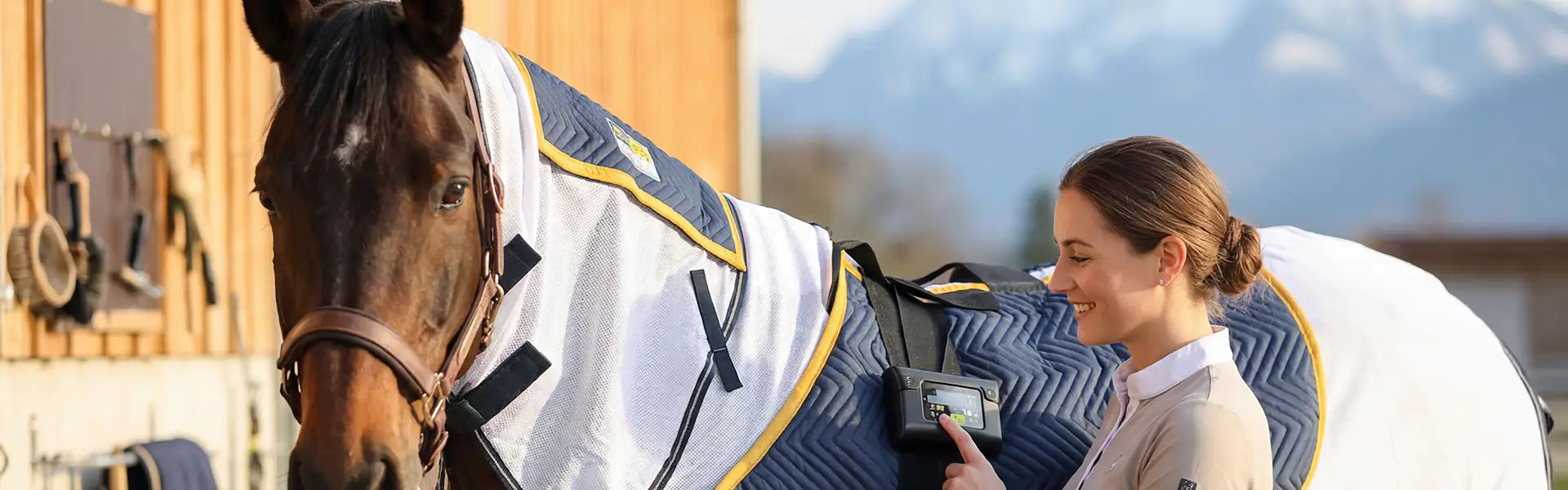 Glückliche Pferdebesitzerin mit einem Pferd, auf dem eine equimag Magnetfelddecke expert Air aufliegt. Die Frau bedient das Steuergerät.